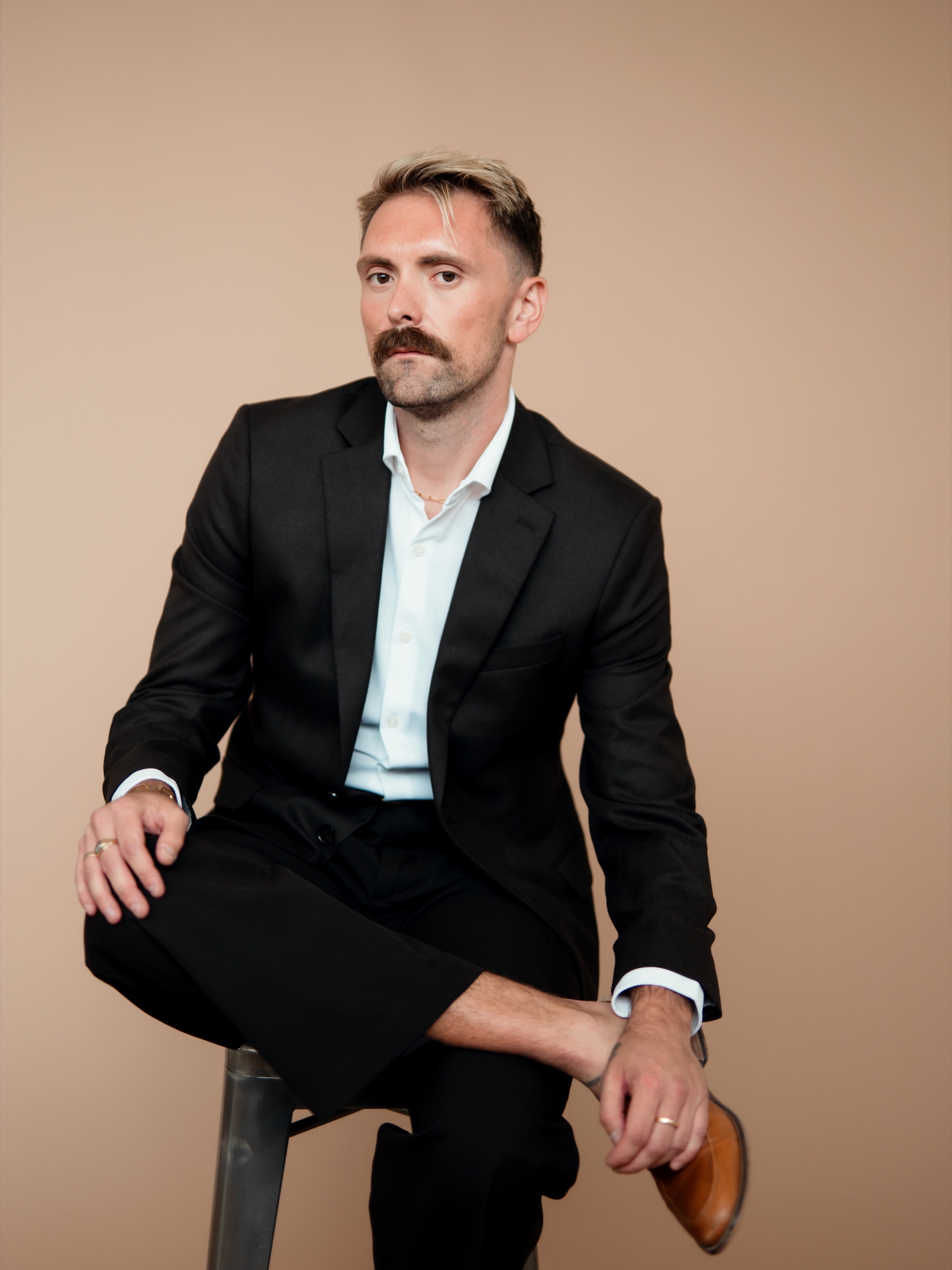 Jordon seated in a black suit with one ankle resting over his knee.