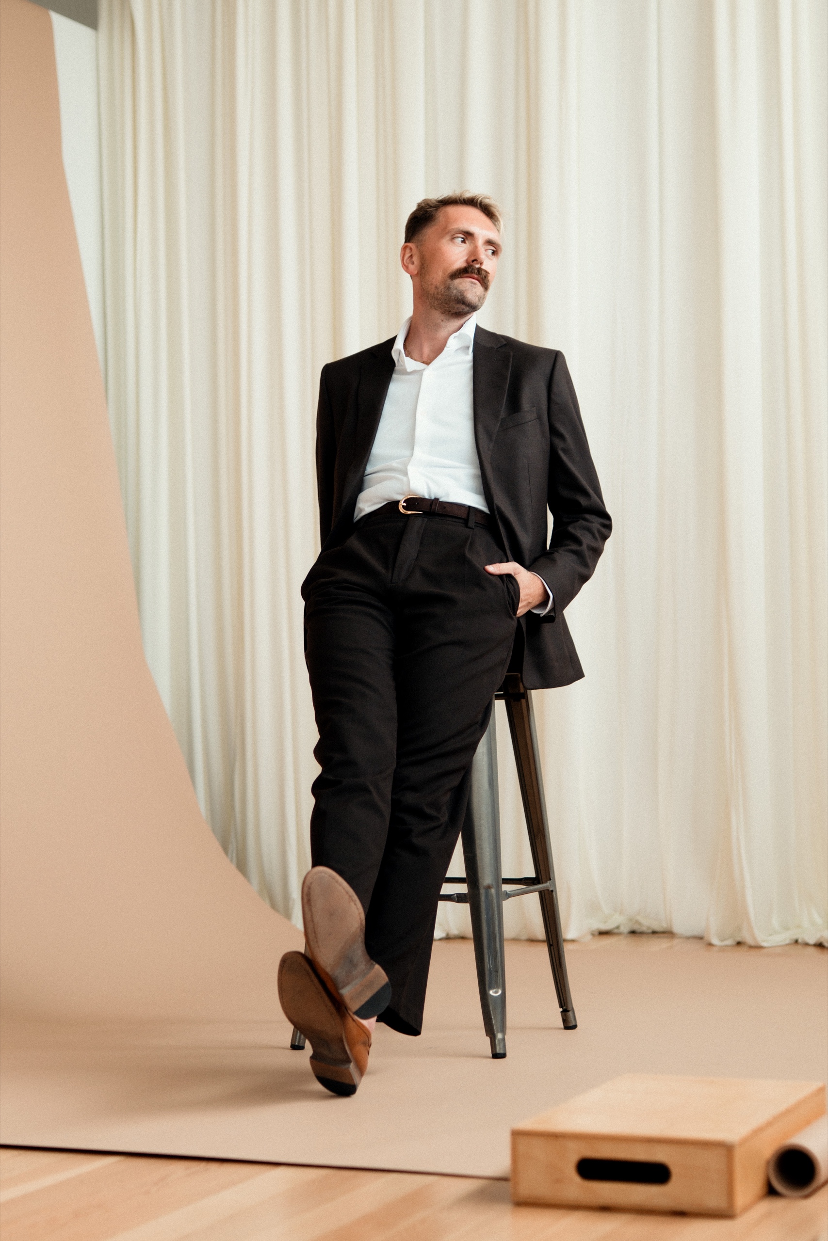 Jordon in a black suit leaning against a tall studio stool.